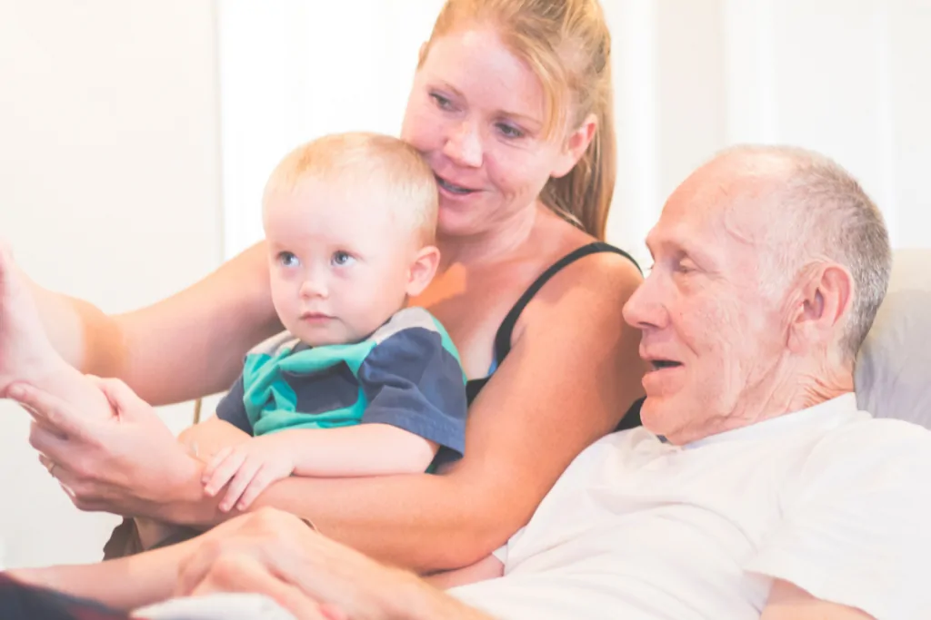 Family with a young child gathered beside an older man resting in bed
