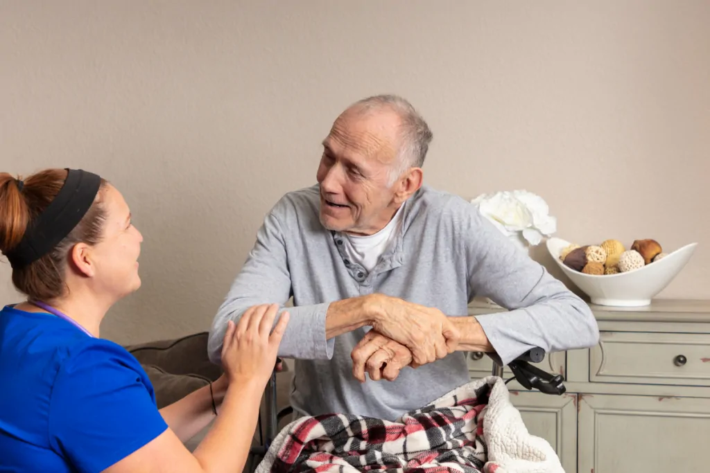 Hospice caregiver kneeling beside an older man in a wheelchair at home